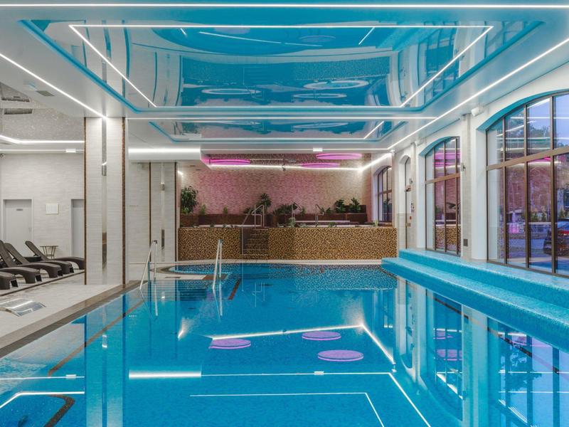 Innenansicht eines modernen Hallenbads mit blauen Wasser, Liegestühlen und dekorativer Deckenbeleuchtung.