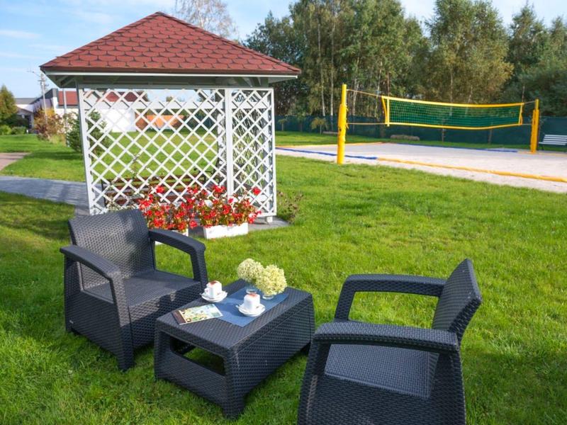 Garten mit zwei schwarzen Stühlen, Tisch mit Teeservice und einer Pergola im Hintergrund neben Volleyballfeld.