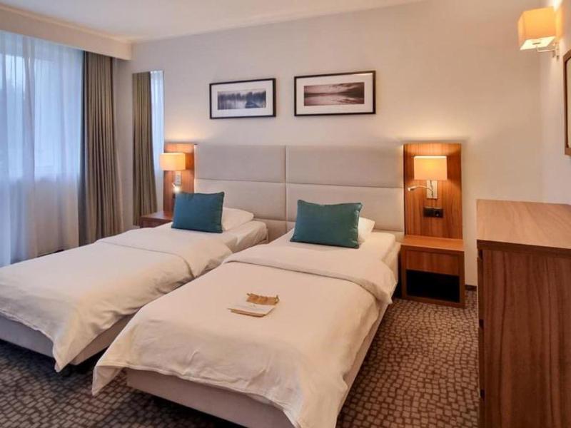 Hotelzimmer mit zwei Einzelbetten, Nachttisch, Schrank und großen Fenstern mit Vorhängen.