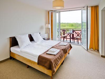 Helles Hotelzimmer mit Doppelbett, brauner Decke und Balkon mit Blick auf grüne Landschaft.