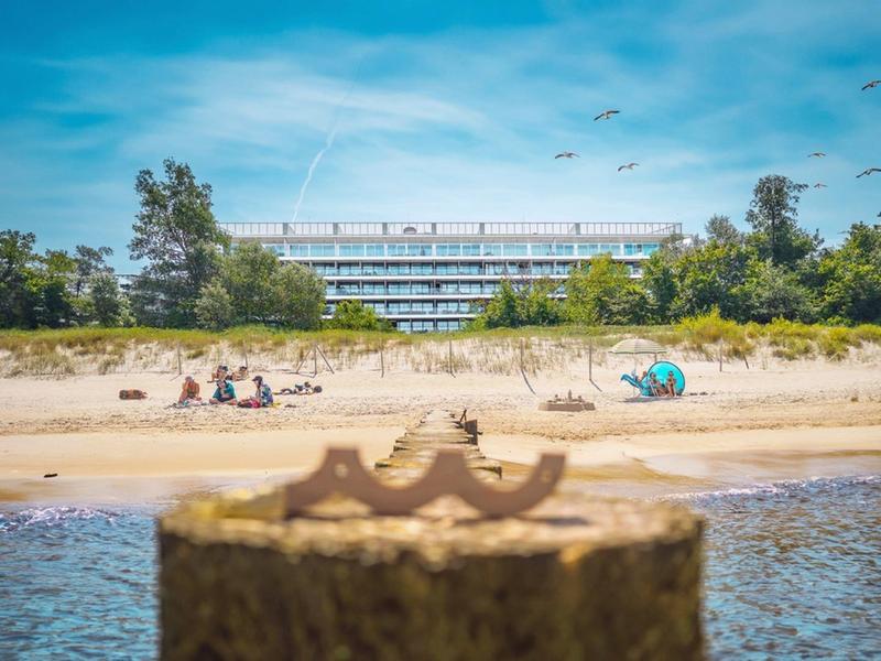 Playa con personas, arena y un edificio de hotel moderno al fondo bajo un cielo azul.
