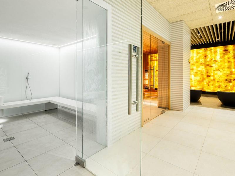 Área de spa moderna con baño de vapor, paredes de vidrio y pared iluminada de piedra amarilla.