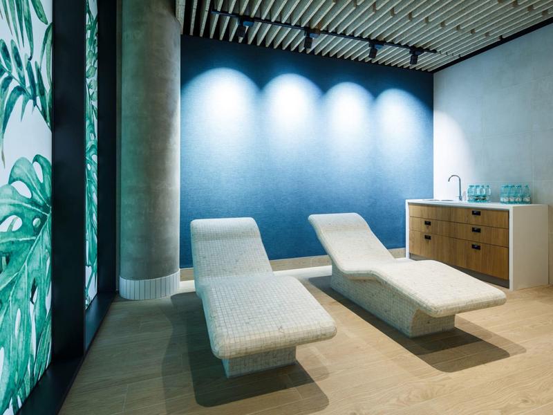 Sala de relajación moderna con tumbonas blancas, pared azul y patrón de plantas en la pared de vidrio.