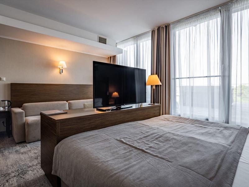 Habitación de hotel moderna con cama, sofá, televisor y grandes ventanas.