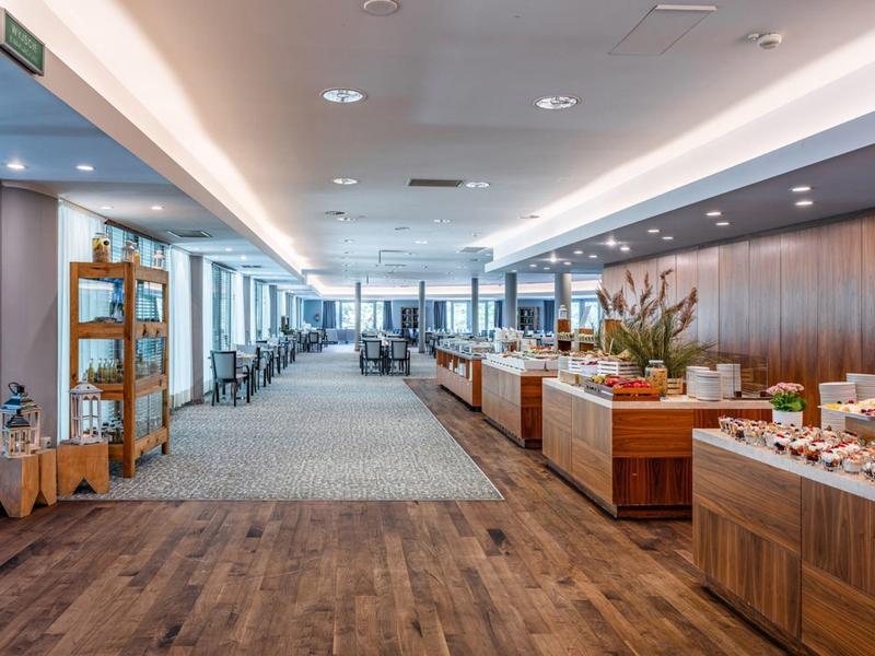 Área de buffet iluminada con suelo de madera y grandes ventanales en un hotel moderno.