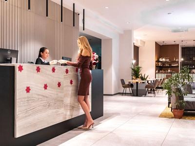 Hotelreceptie met twee vrouwen, een staat bij de balie en de ander zit erachter, modern interieur met planten.