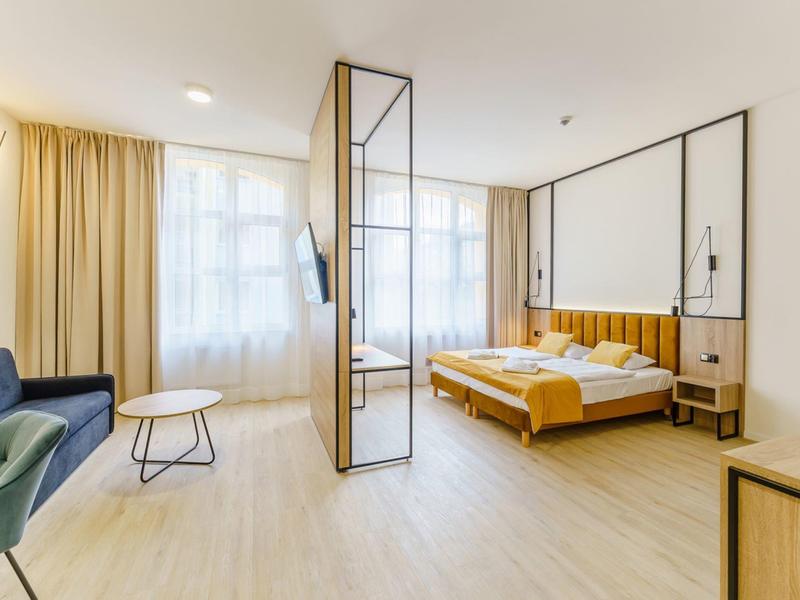 Helles Hotelzimmer mit Doppelbett, Sofa, Vorhängen und einem Raumteiler aus Glas und Metall.