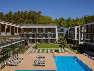Moderner Hotelkomplex mit Pool, Liegestühlen und Gartenfläche vor bewaldetem Hintergrund.