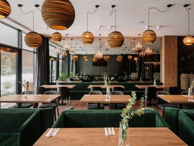 Nowoczesna restauracja z drewnianymi stołami, zielonymi welurowymi krzesłami i okrągłymi drewnianymi lampami sufitowymi.