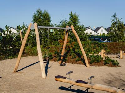 Huśtawka i bujak na piaszczystym placu zabaw z domami i krzewami w tle