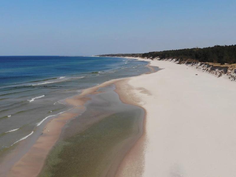Rozległa biała piaszczysta plaża z spokojnym niebieskim morzem i zalesionym tłem pod niebieskim niebem.