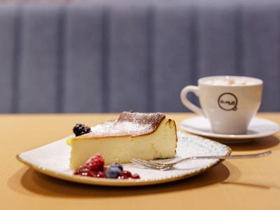 Kousek sýrového dortu na bílém talíři s bobulemi, vedle šálek kávy na podšálku.