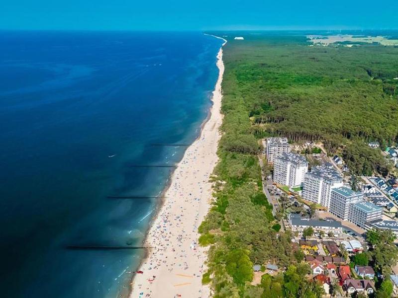 Letecký pohled na dlouhou písčitou pláž s modrým mořem, přilehlým lesem a budovami.