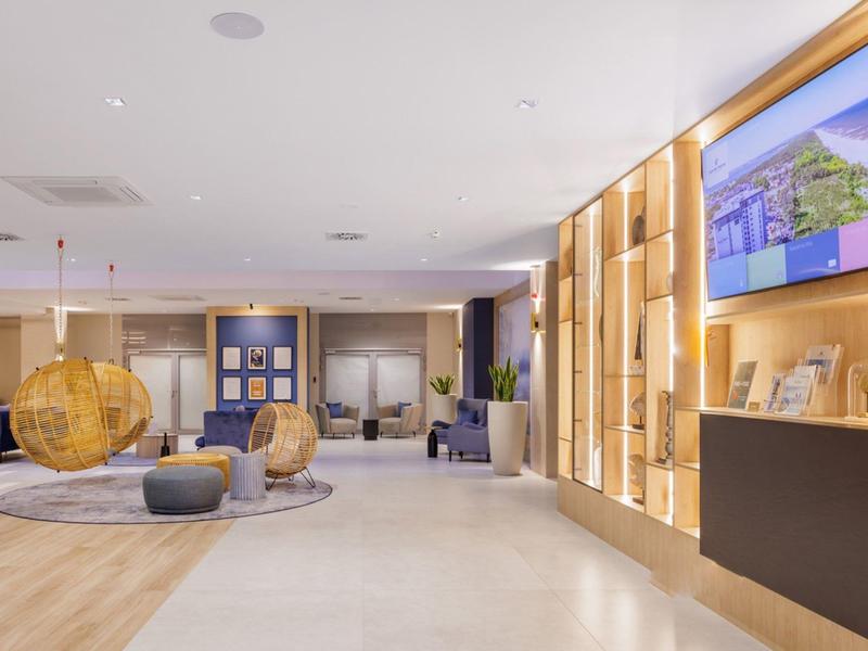 Moderní hotelová lobby s posezením, dřevěnými panely a velkou televizní obrazovkou.