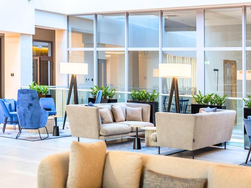 Světlá hotelová hala s moderními pohovkami, křesly a dekorativními rostlinami za denního světla.