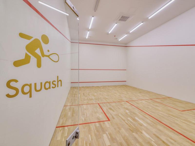 Prázdný squashový kurt se světlou dřevěnou podlahou a bílými stěnami se symbolem squashu.