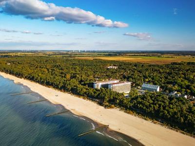 Strand mit klarem Wasser, Sand und bewaldetem Gebiet mit Gebäuden im Hintergrund unter blauem Himmel.