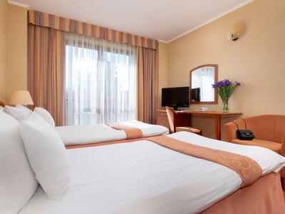 Helles Hotelzimmer mit zwei Betten, einem Schreibtisch, Stuhl und Fenster mit Vorhängen.