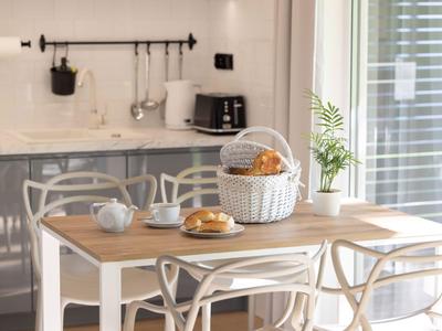 Gemütlicher Essbereich mit Holz- und weißen Stühlen, Korb mit Brötchen und einer Pflanze nahe eines Fensters.