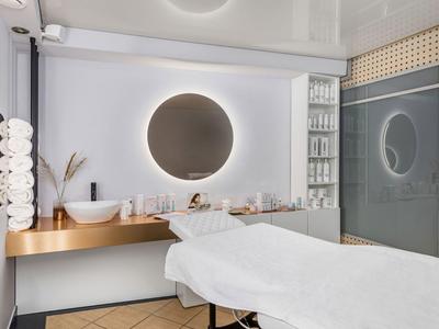 Behandlungsraum mit Liege, rundem beleuchtetem Spiegel und Regalen mit Kosmetikprodukten.