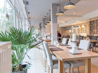 Helles Restaurant mit Holztischen, modernen Stühlen und großen Fenstern mit Blick auf Pflanzen.