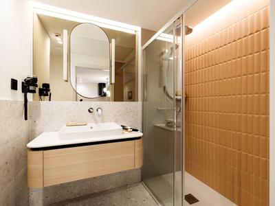 Moderní hotelová koupelna se umyvadlem, velkým zrcadlem a béžovou sprchovou stěnou.