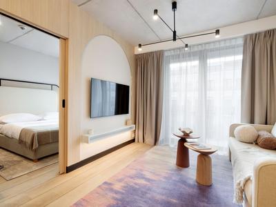 Světý hotelový pokoj s obývacím koutem, TV na dřevěné stěně a sousední ložnicí s velkou postelí.