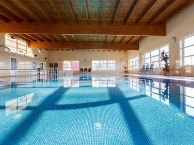 Piscina interna luminosa con acqua blu, soffitto in legno e grandi finestre.