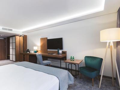 Area moderna di camera d'hotel con letto, scrivania, TV e poltrona sotto luce soffusa.