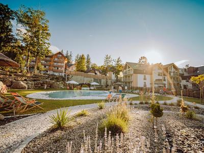 Complesso alberghiero con piscina, circondato da alberi e montagne sotto un cielo limpido.