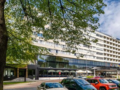 Grande edificio di hotel a più piani con parcheggio davanti, incorniciato da rami di alberi.