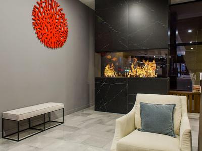 Area lounge moderna dell'hotel con camino, poltrona beige, cuscino blu e decorazione murale rossa.