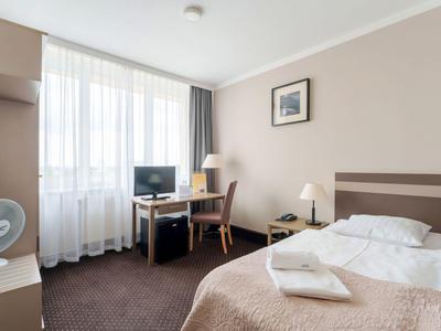 Camera d'hotel luminosa e moderna con finestra ampia, letto singolo, scrivania e TV.