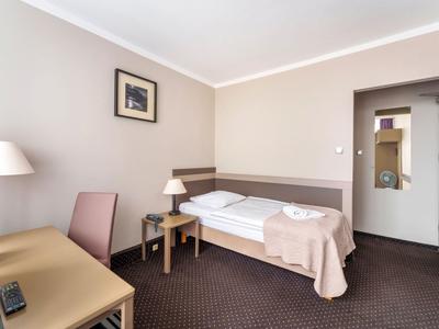 Una semplice camera d'hotel con un letto singolo, scrivania, sedia e due comodini su pareti neutre.