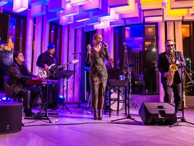 Band dal vivo con cantante, tastierista, chitarrista e sassofonista in una stanza moderna illuminata a colori.