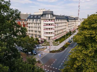 Vista de un edificio de hotel de varios pisos en una intersección, rodeado de árboles.