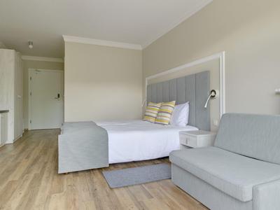 Habitación de hotel luminosa con cama doble, sofá gris y suelo de madera.