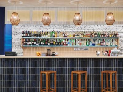 Espace bar d'hôtel moderne avec tabourets, étagères remplies de bouteilles et mur en briques clair.