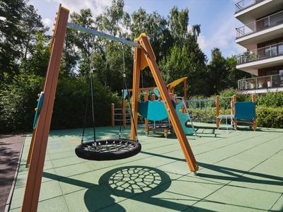 Aire de jeux avec une balançoire nid et une structure d'escalade à côté d'un immeuble résidentiel et des arbres sous un ciel bleu.