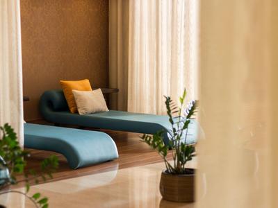 Interno moderno di un hotel con due chaise longue blu e piante vicino a una grande finestra con tende.