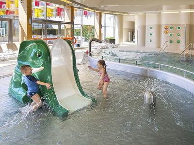 Bambini che giocano su uno scivolo verde in acqua bassa di una piscina coperta con giochi d'acqua.