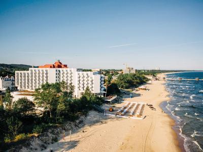 Hotel an einem breiten Sandstrand mit Blick auf das blaue Meer und bewaldete Hügel.