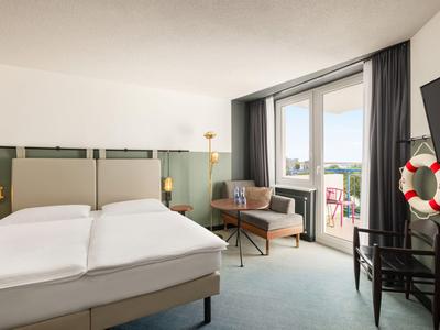 Helles Hotelzimmer mit Doppelbett, Sessel, Balkon und Fernseher an der Wand.