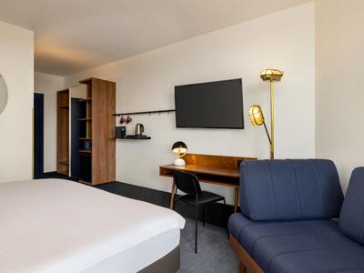 Hotelzimmer mit Doppelbett, Arbeitsplatz, Fernseher und blauer Couch in schlichtem Design.