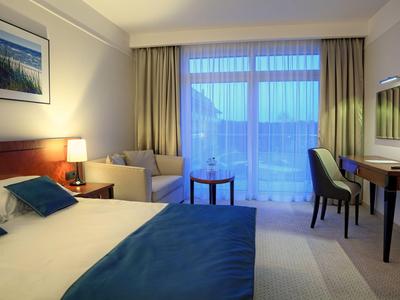 Habitación de hotel luminosa con cama doble, área de estar y ventana grande con vista a las montañas.