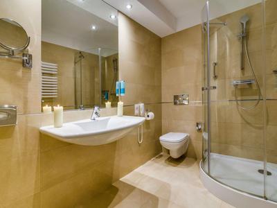 Baño de hotel moderno con ducha de vidrio, inodoro y lavabo en tonos beige cálidos.