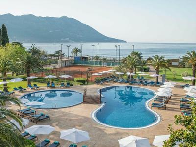 Hotelpool mit Liegen und Sonnenschirmen am Meer, umgeben von Palmen und Bergen im Hintergrund.