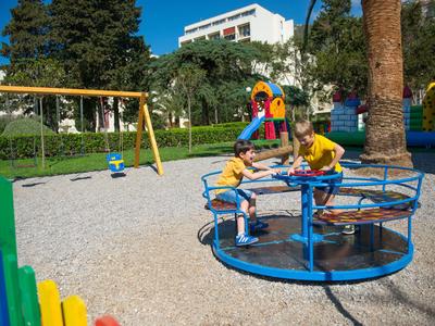 Kinder spielen auf einem Karussell in einem sonnigen Spielplatz im Hotelgarten.