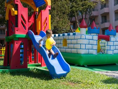 Kind rutscht eine blaue Rutsche an einem buntfarbenen Spielplatz im Hotelgarten hinunter.