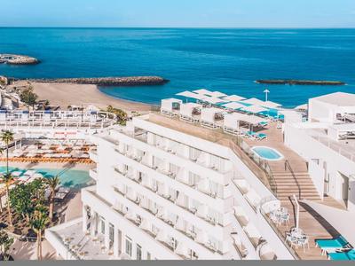 Großes weißes Hotel am Meer mit mehreren Pools, Terrassen und blauem Himmel über der Küste.